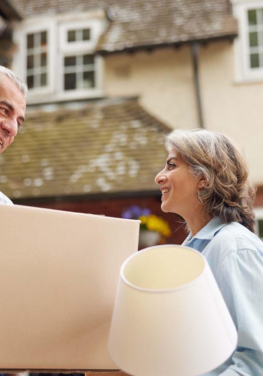 Mature Couple Carrying Boxes On Moving Day In Front Of Dream Home